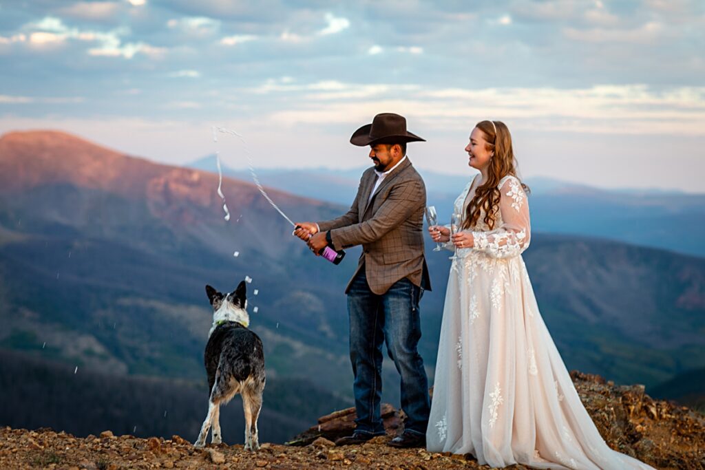 cowboy groom opening a bottle of champagne while bride and dog observe